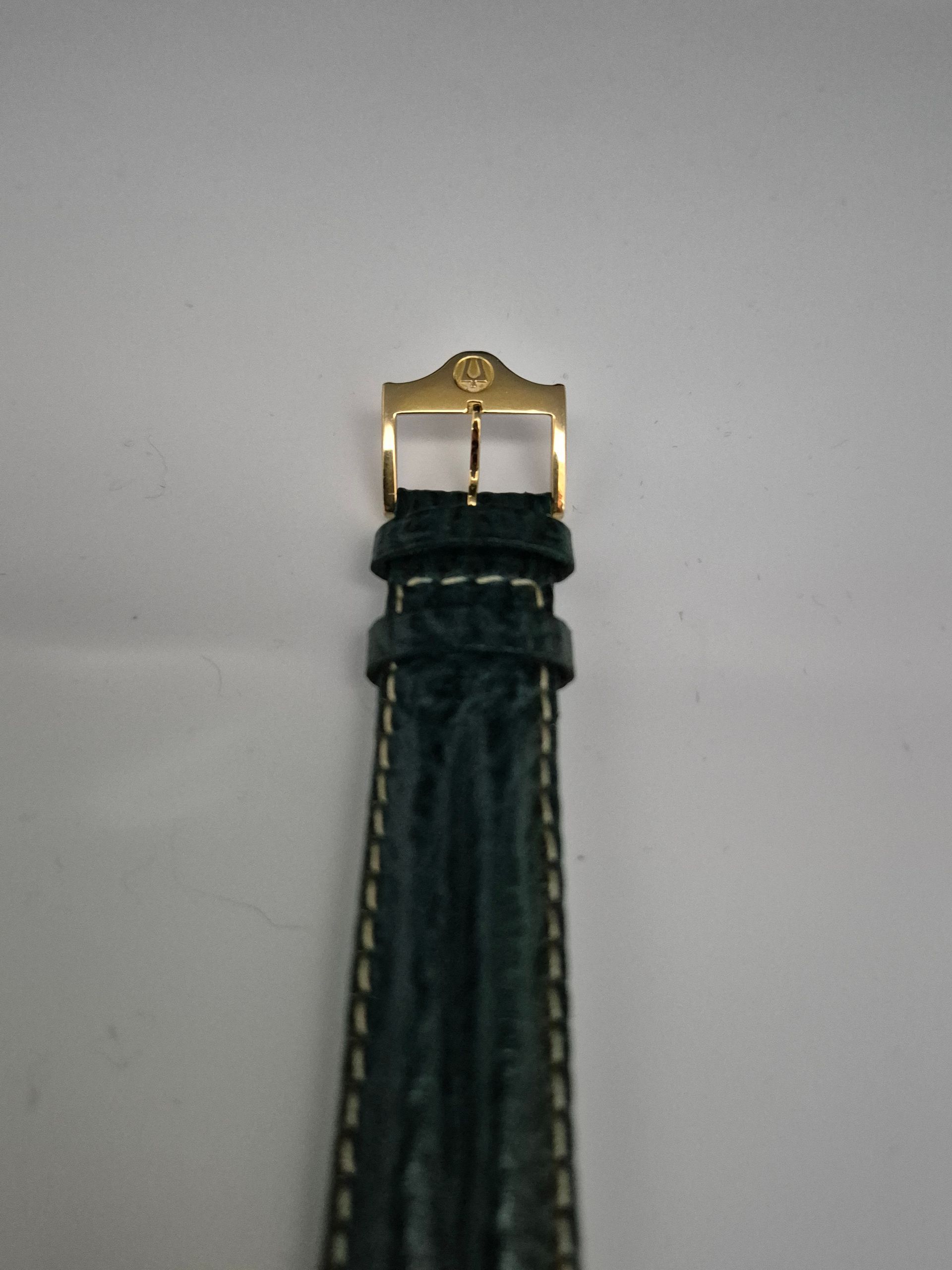 Close-up of a dark green leather watch strap with a gold buckle against a light background