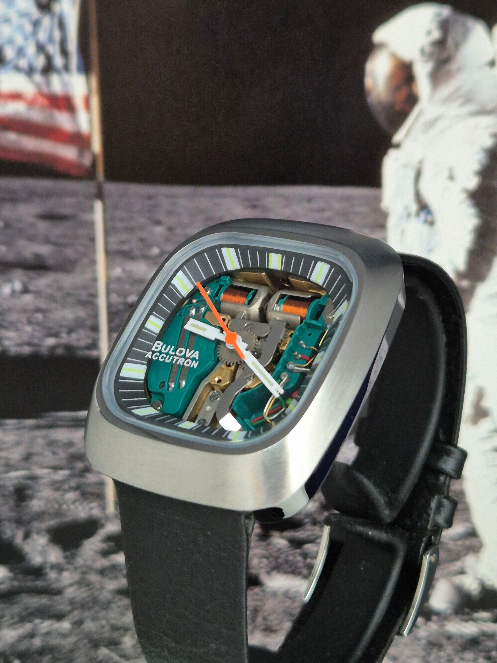Square silver watch with a teal open-work face and black strap on a lunar surface background