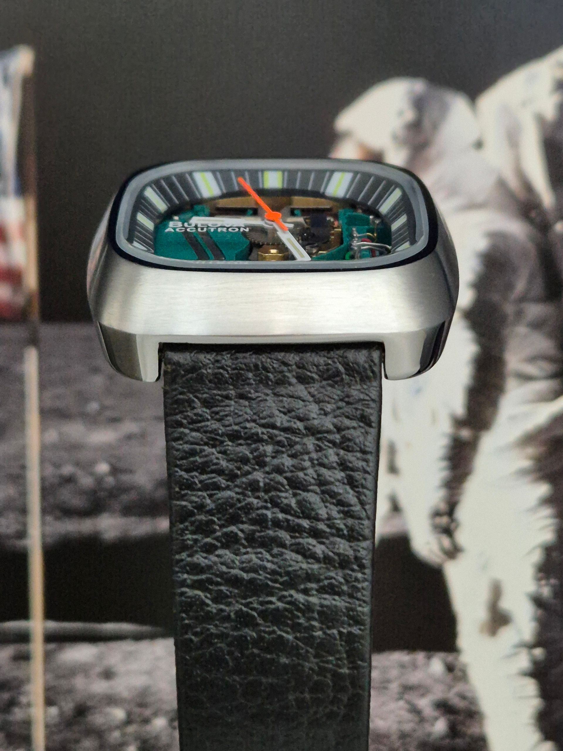 Close-up of a rectangular silver wristwatch with a black textured strap against a blurred astronaut backdrop