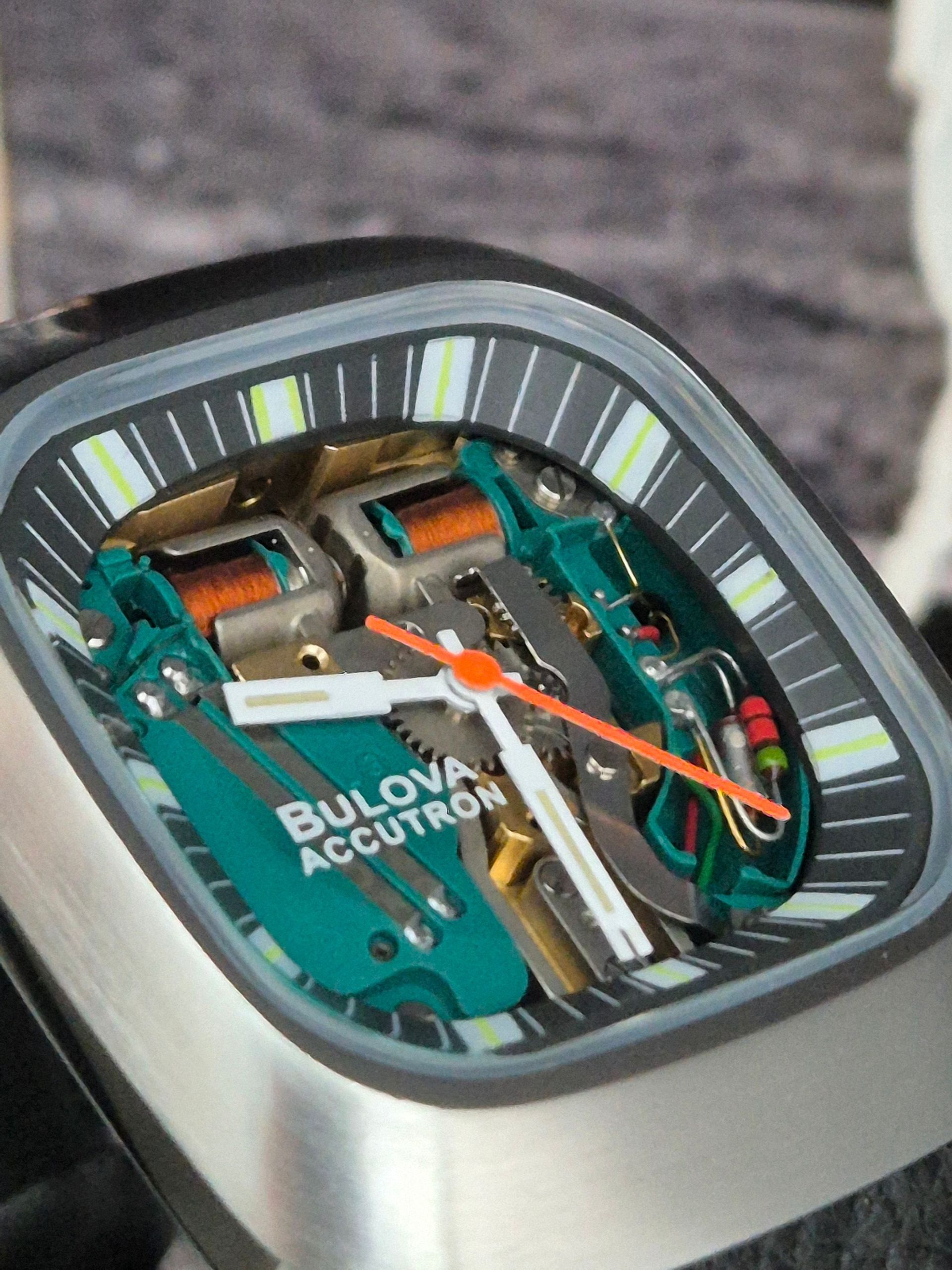 Close-up of a futuristic square watch with a green and silver dial and orange hands on a dark surface