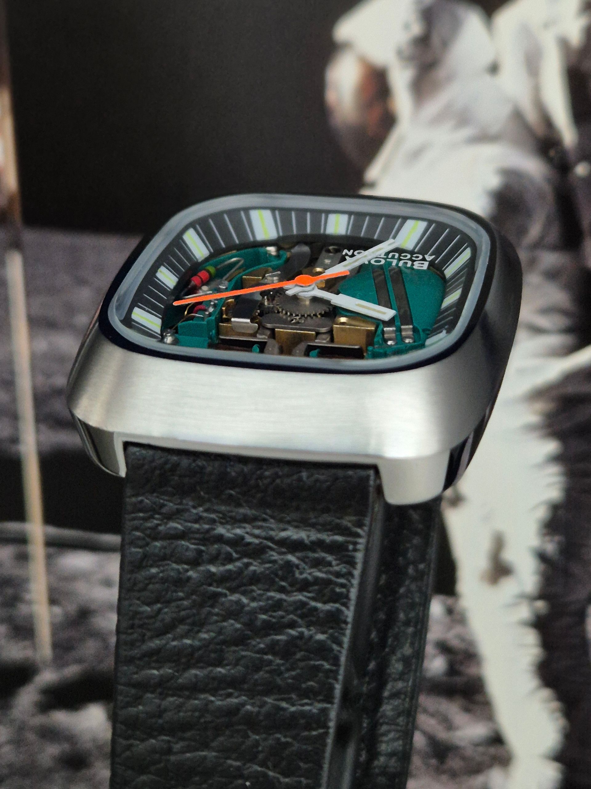 Square silver watch with exposed teal and orange dial on a black leather strap, against a blurred space backdrop
