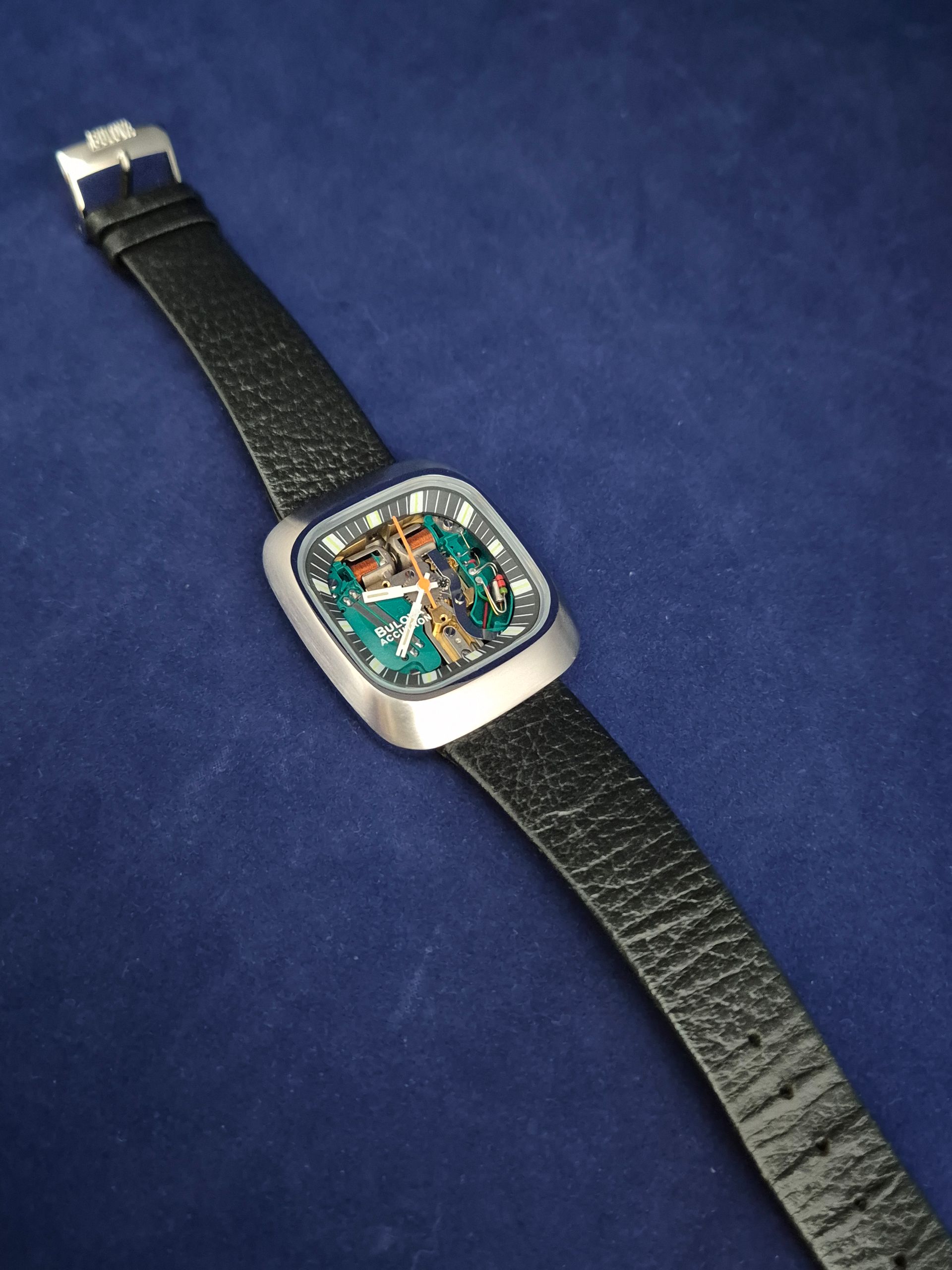 Silver wristwatch with a blue-green face and black textured band on a blue background