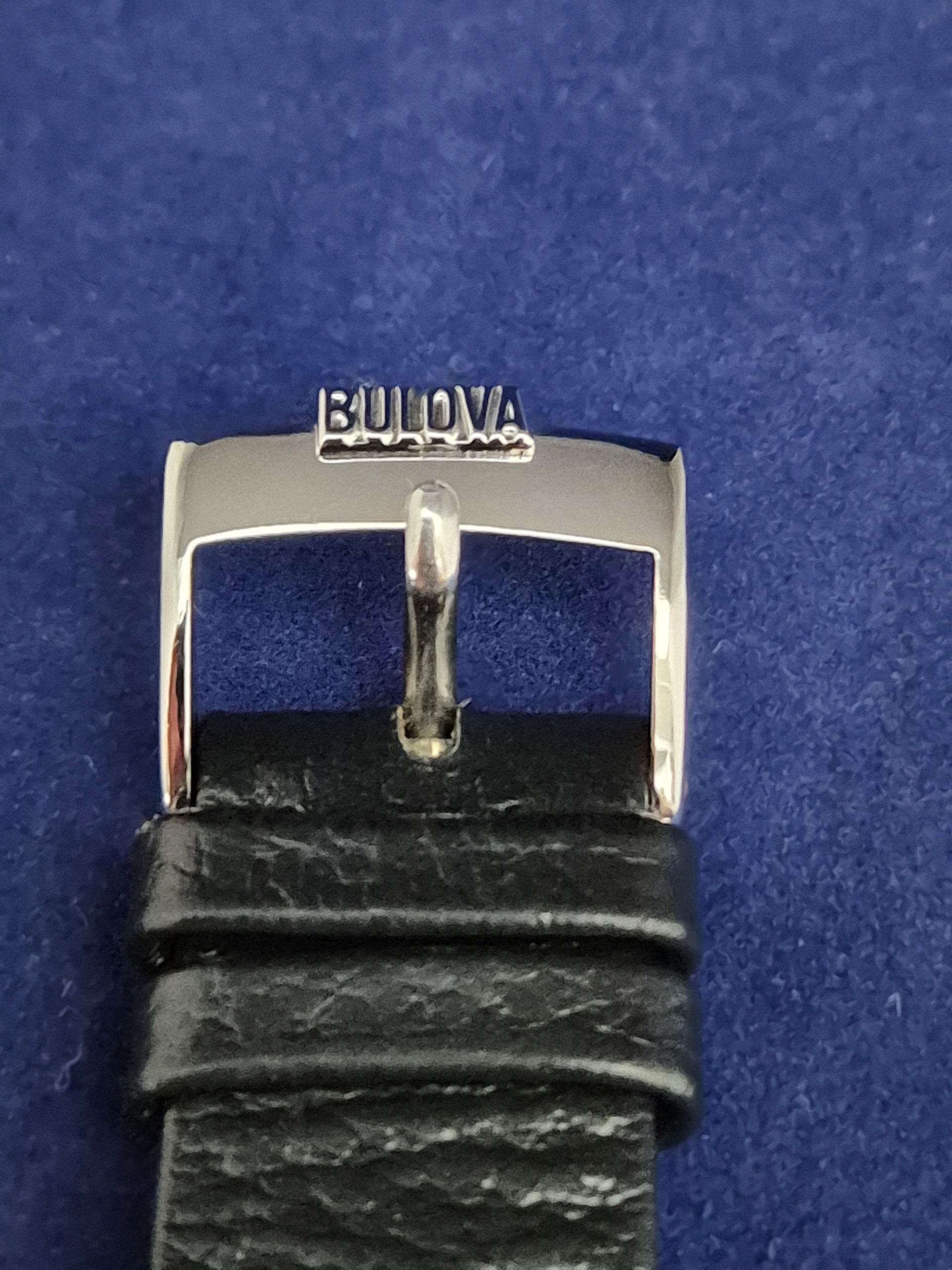 Close-up of a Bulova watch clasp on a black leather strap against a blue background