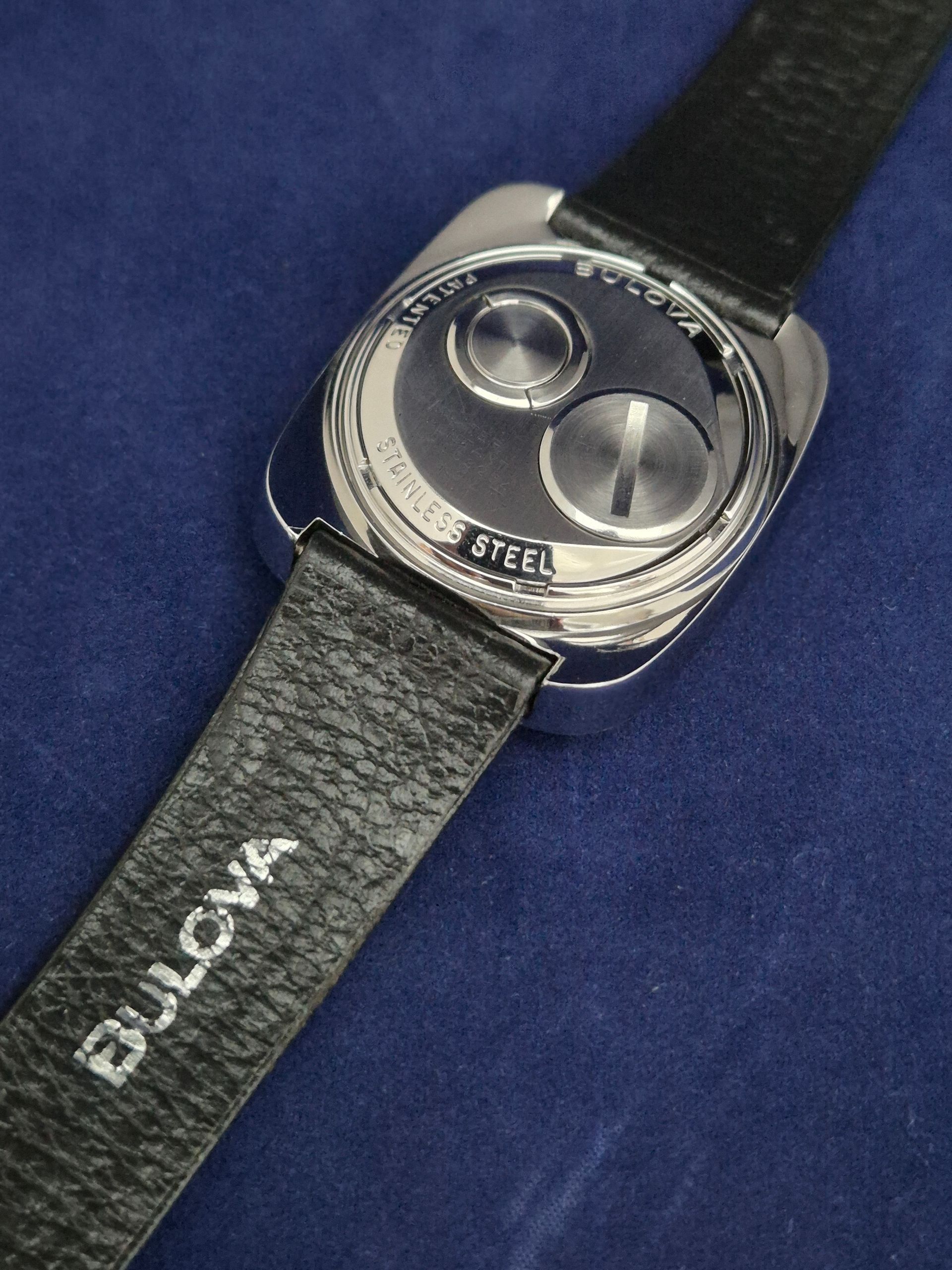 Black wristwatch with rounded silver face and black strap on a blue background.