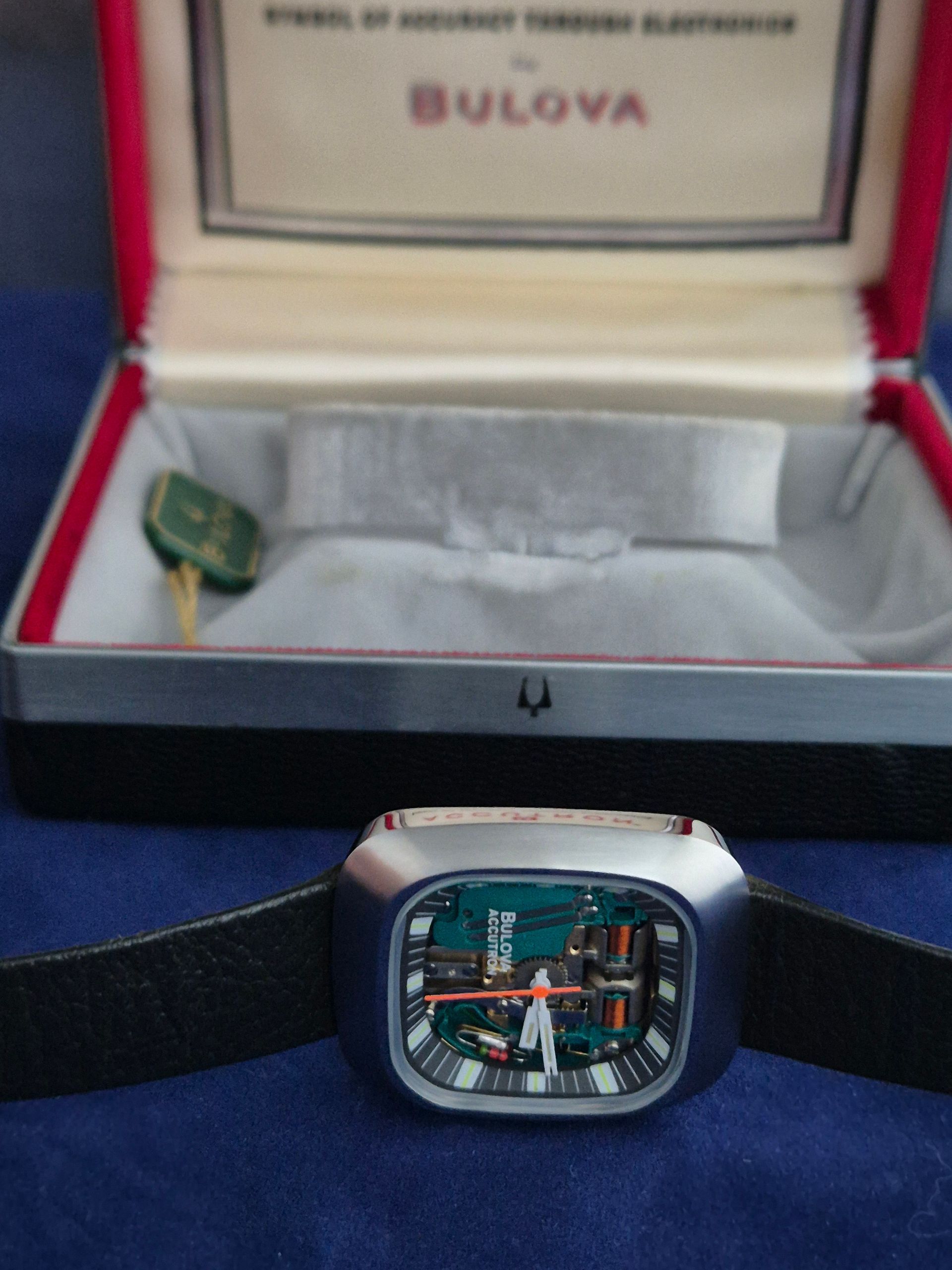 Bulova watch in black case with green tag, open presentation box behind it on blue cloth.