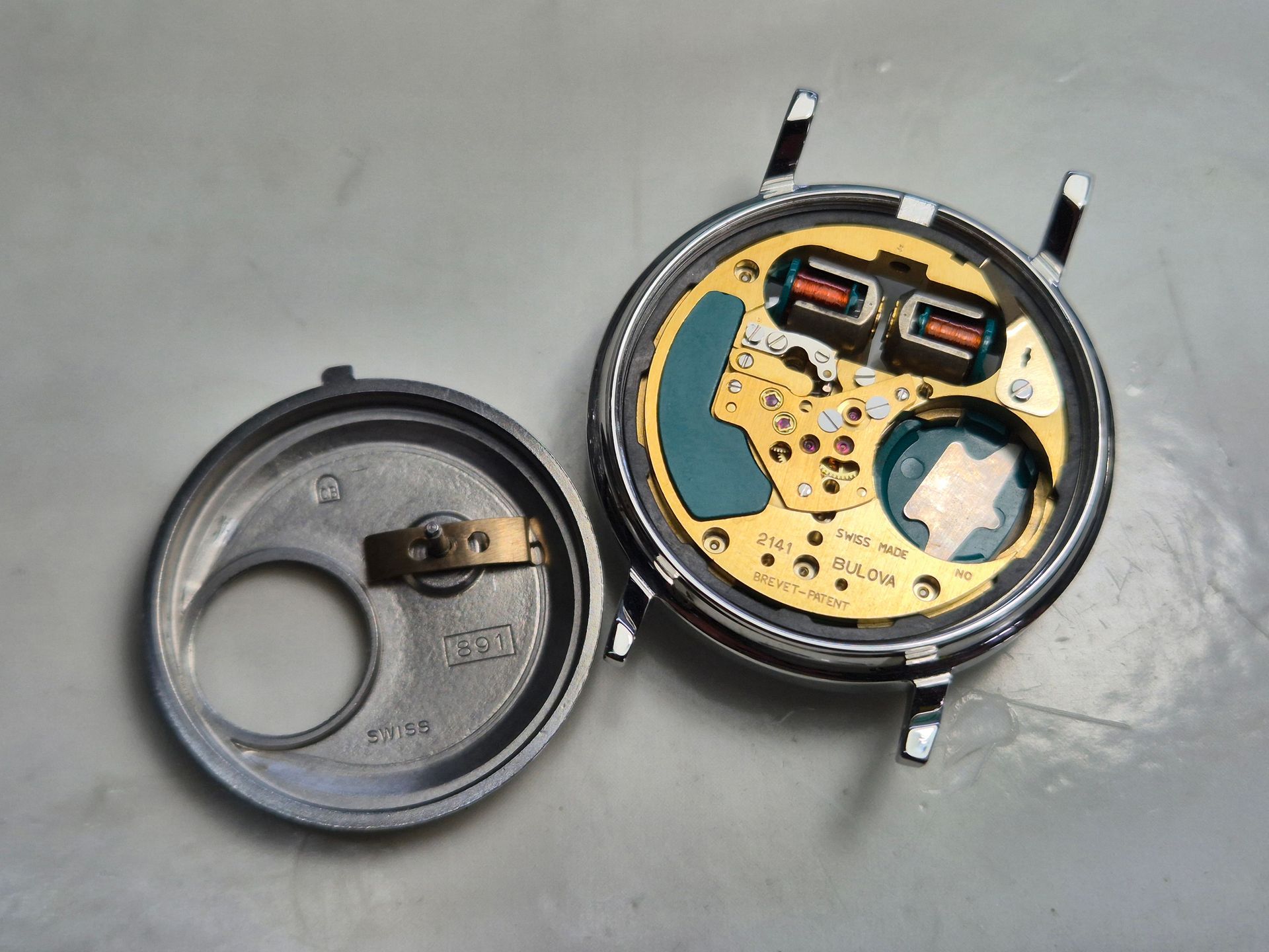 Open wristwatch case showing movement and removed back cover on a gray surface