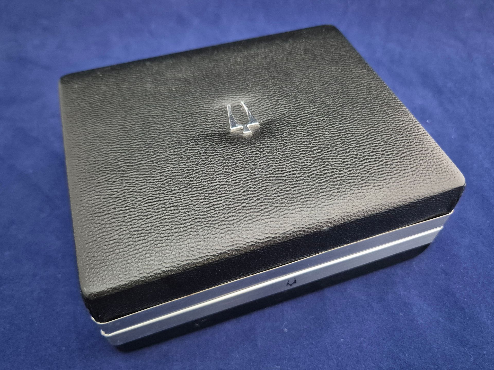 Black textured jewelry box with silver trim and a small silver emblem on top, resting on a blue surface
