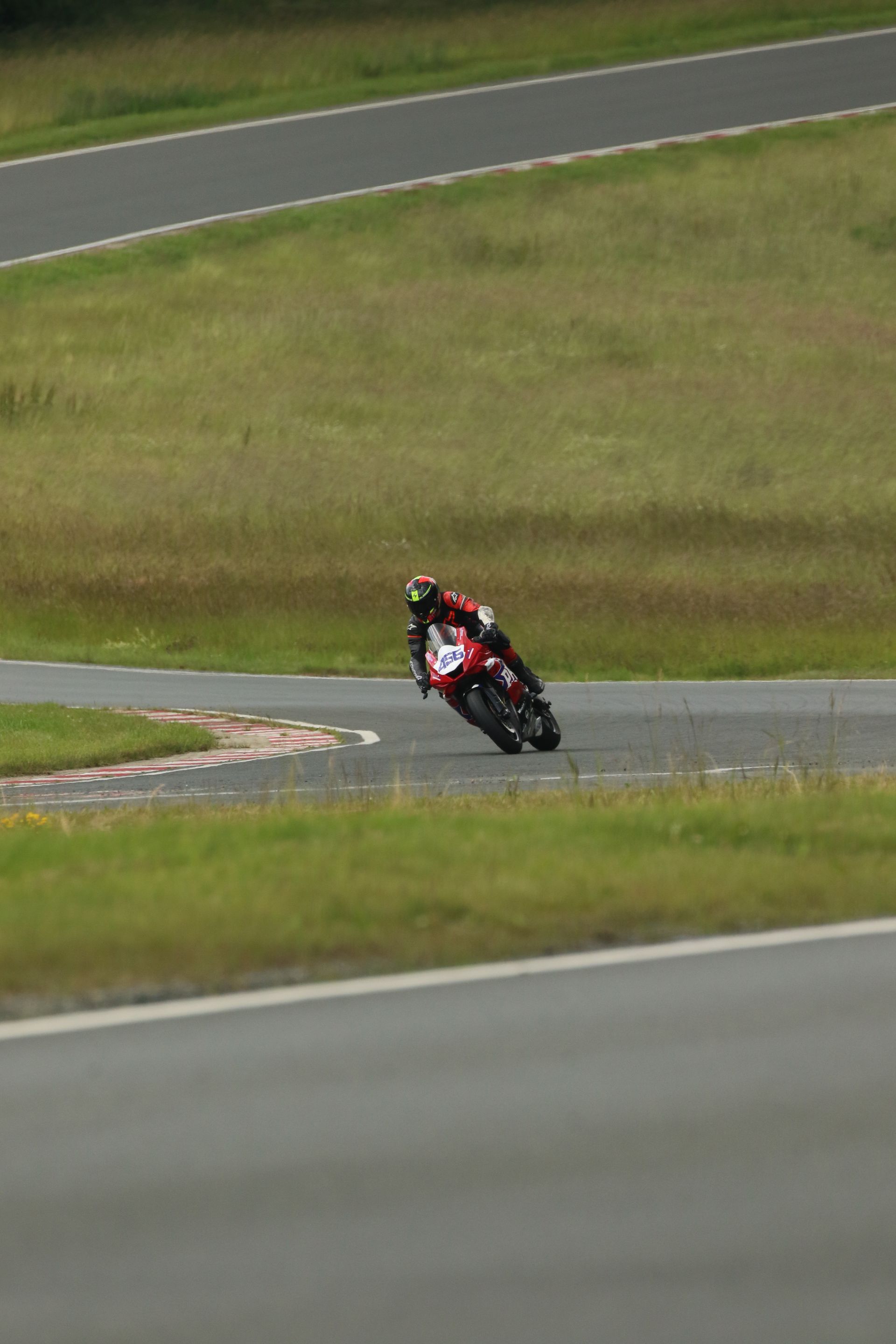 Motorcoureur leunt in een bocht op een circuit. Rood-zwarte motorfiets; groene gras- en asfaltachtergrond.