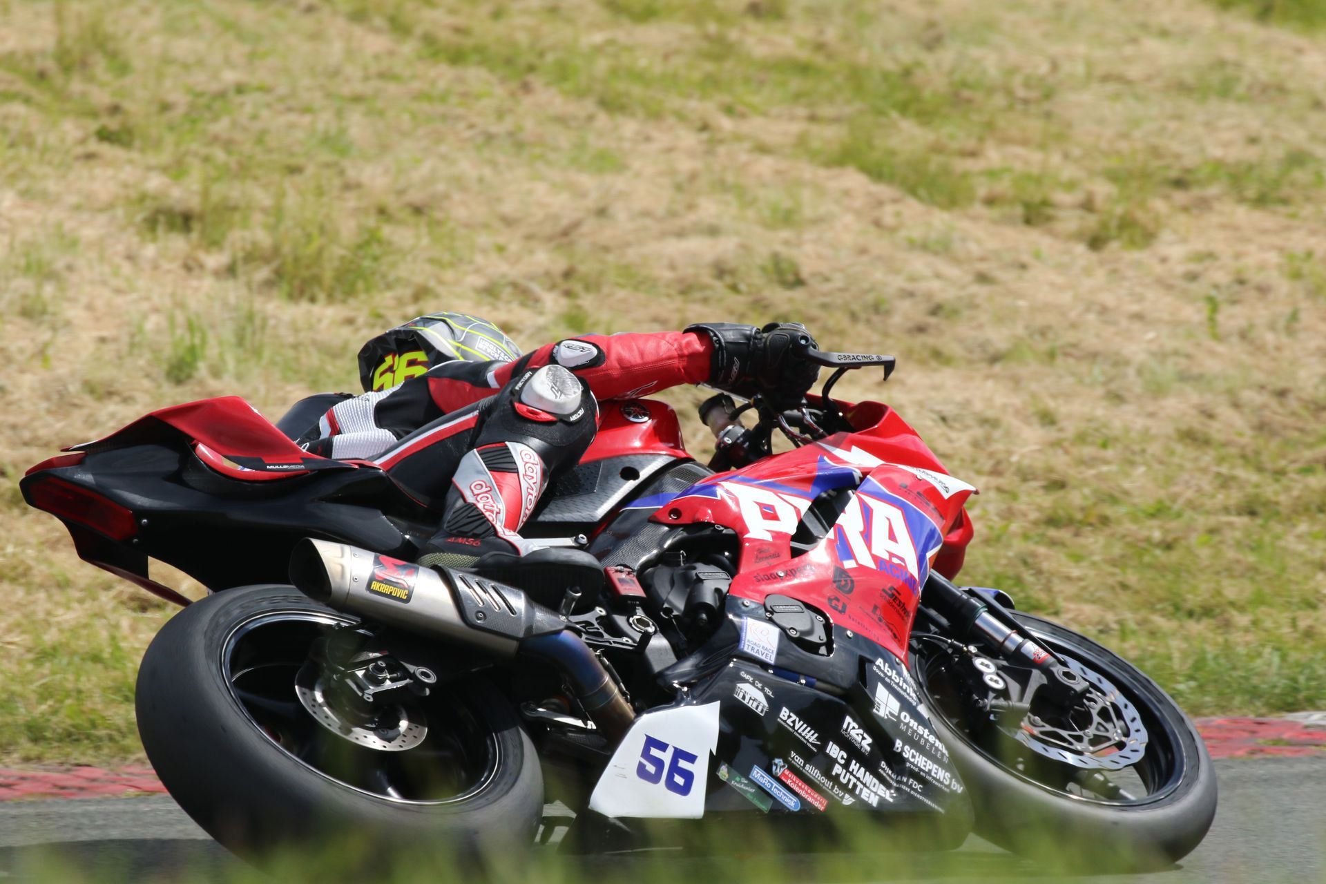 Motorcoureur leunt in een bocht op een circuit, gekleed in rood-zwarte kleding. Zijn motor is rood-zwart en heeft logo's van sponsoren.