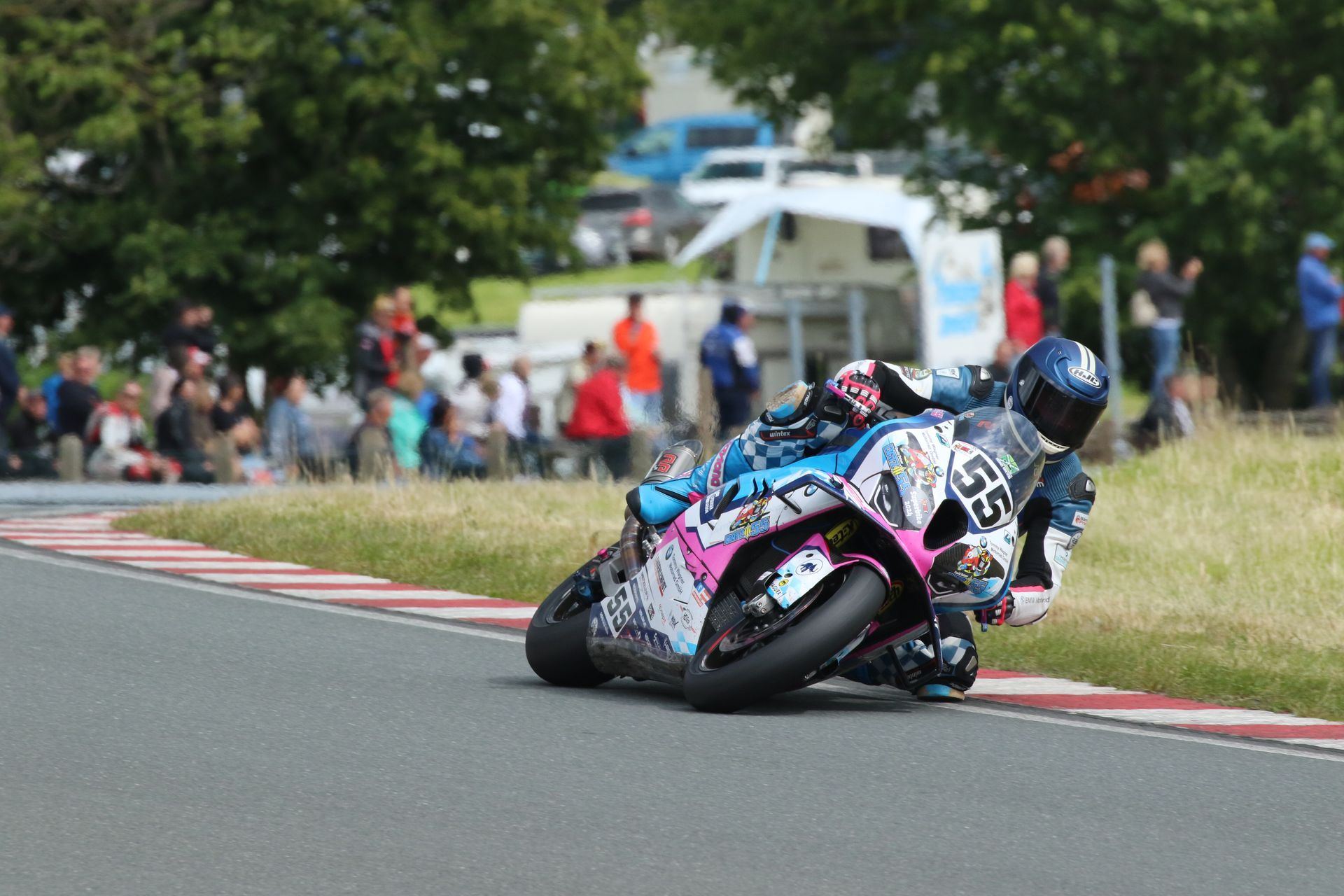 Een motorrijder in zwart en roze kleding leunt in een bocht op een circuit, nummer 55 is zichtbaar, met toeschouwers op de achtergrond.