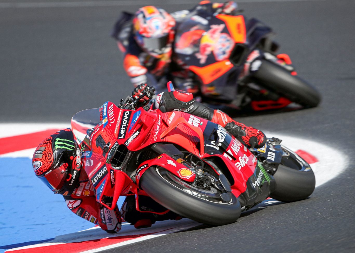 Twee MotoGP-motoren racen op een circuit. De leidende rode Ducati helt scherp een bocht in, gevolgd door een wazig oranje KTM.