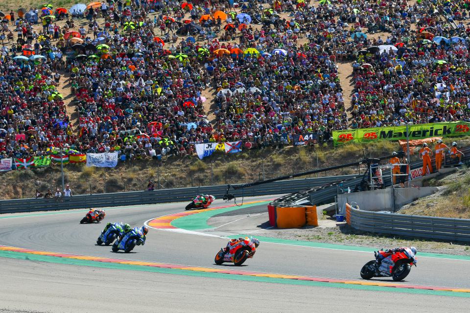 Motorrace op een circuit, met meerdere coureurs die met hoge snelheid door een bocht racen. Een grote menigte kijkt toe vanaf de heuvel.
