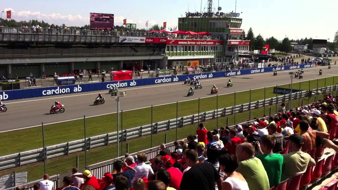 Motorrace aan de gang op een circuit. Een menigte toeschouwers kijkt vanaf de voorgrond toe, met coureurs op de baan in de verte.