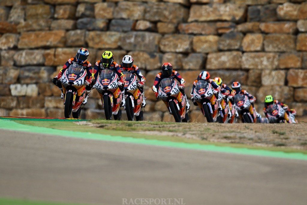 Motorcoureurs in kleurrijke kleding racen over een circuit bij een stenen muur. Verschillende motoren leunen een bocht in.