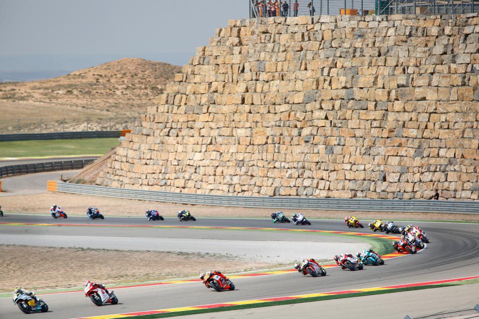 Motorrace op een circuit, met coureurs die bochten nemen, een grote stenen muur op de achtergrond en toeschouwers bovenaan.