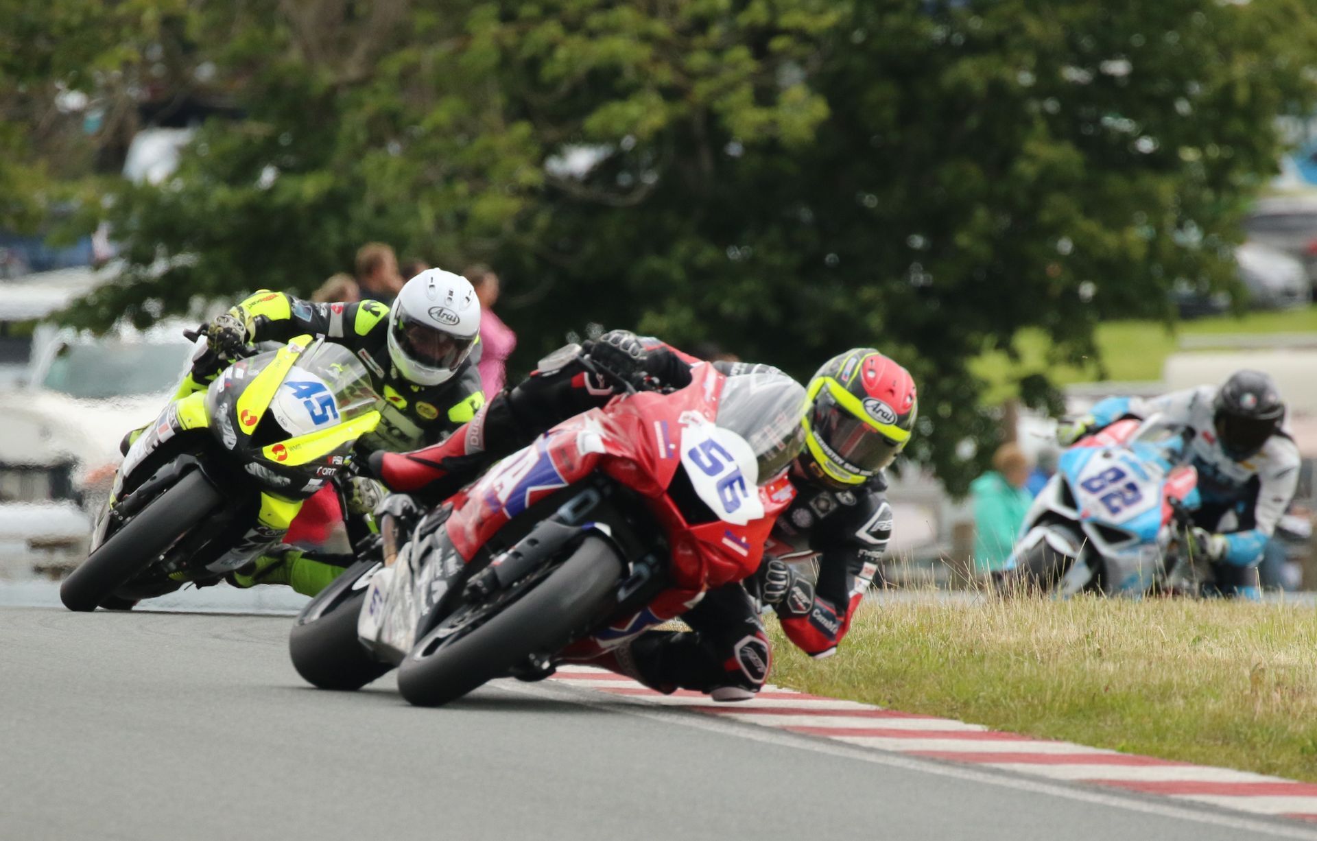Motorcoureurs leunen in een bocht op een circuit. Drie coureurs in kleurrijke uitrusting strijden, met een zichtbaar toeschouwersgebied.