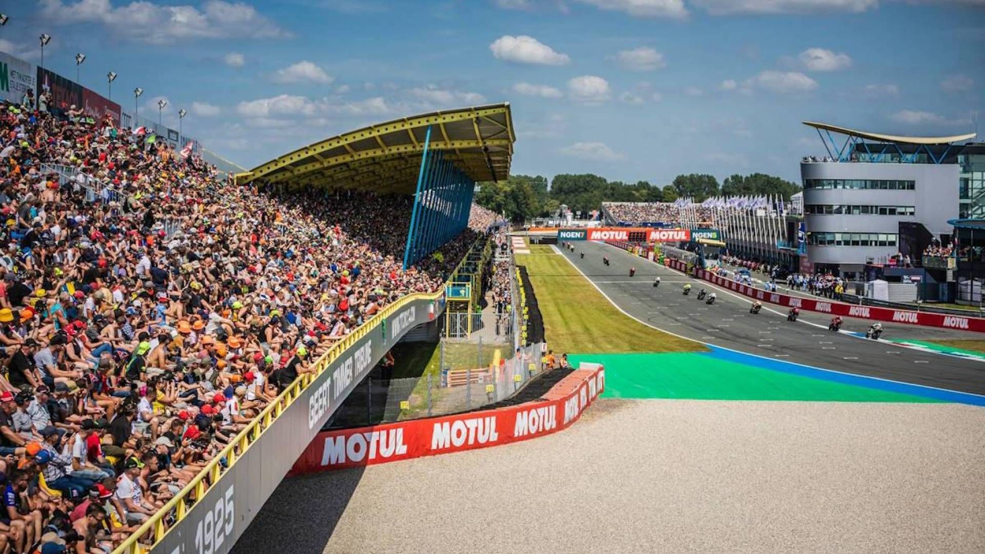 Een druk racecircuit waar een motorrace gaande is, gezien vanaf de tribune in het stadion, met de kleuren blauw, groen en geel.