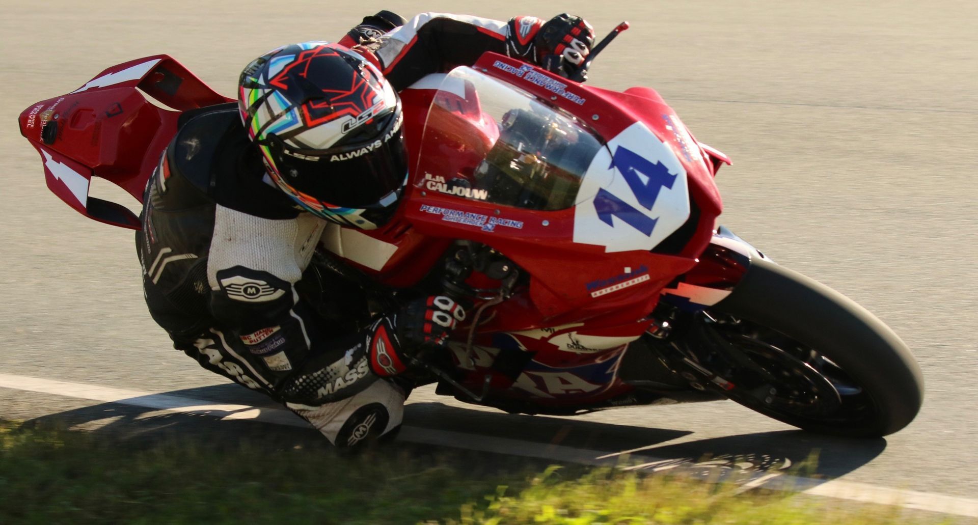 Motorcoureur in rood-wit pak buigt zich voorover op een circuit, helm naar beneden, motor schuin, nummer 14 op de voorkant.