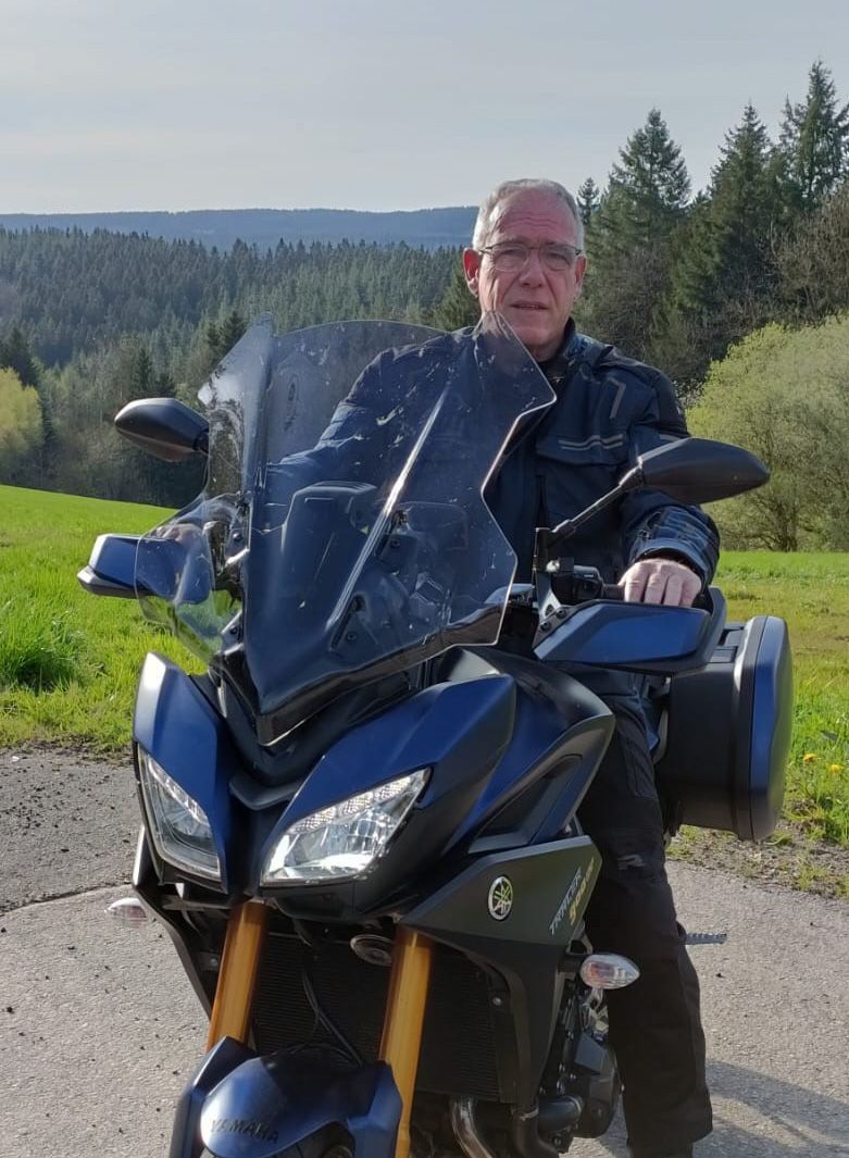 Een man op een blauwe motor poseert buiten op een verharde weg. Hij draagt een donker jack. Op de achtergrond is een bos te zien.