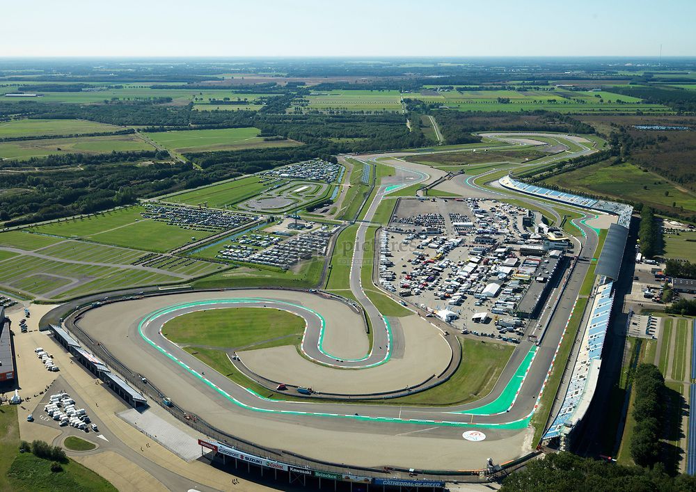 Luchtfoto van een racebaan met een helderblauw en groen baanoppervlak, omgeven door infrastructuur en groene velden onder een blauwe lucht.