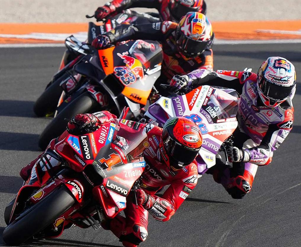 Motorcoureurs in rood, oranje en zwart racen dicht op elkaar op een circuit. De coureurs leunen schuin in een bocht.