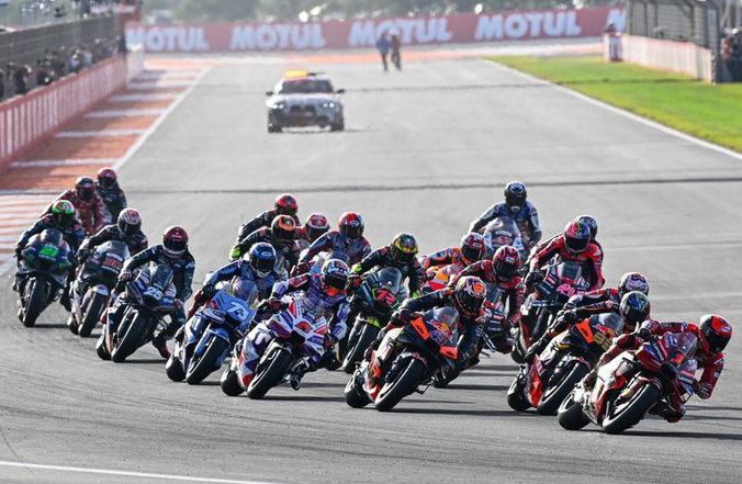 Motorcoureurs racen over een circuit, oranje en zwarte motoren prominent voorop. In de verte zijn een auto en mensen te zien.