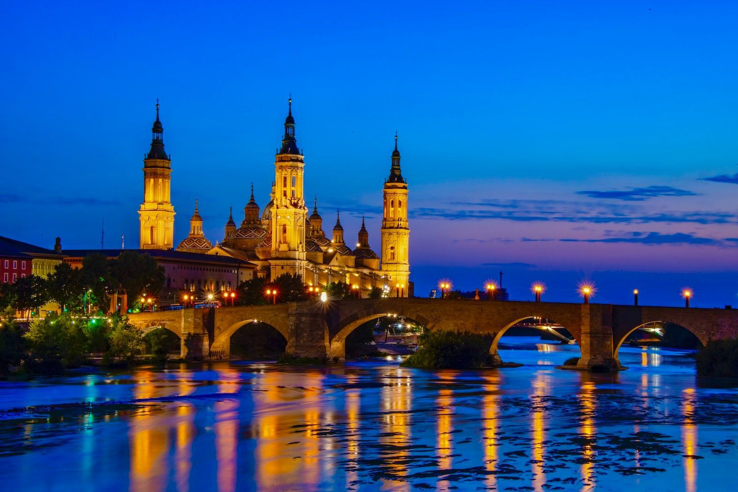 Nachtzicht op de verlichte Basilica del Pilar in Zaragoza, Spanje, met een weerspiegeling in de rivier. Een brug overspant het water.