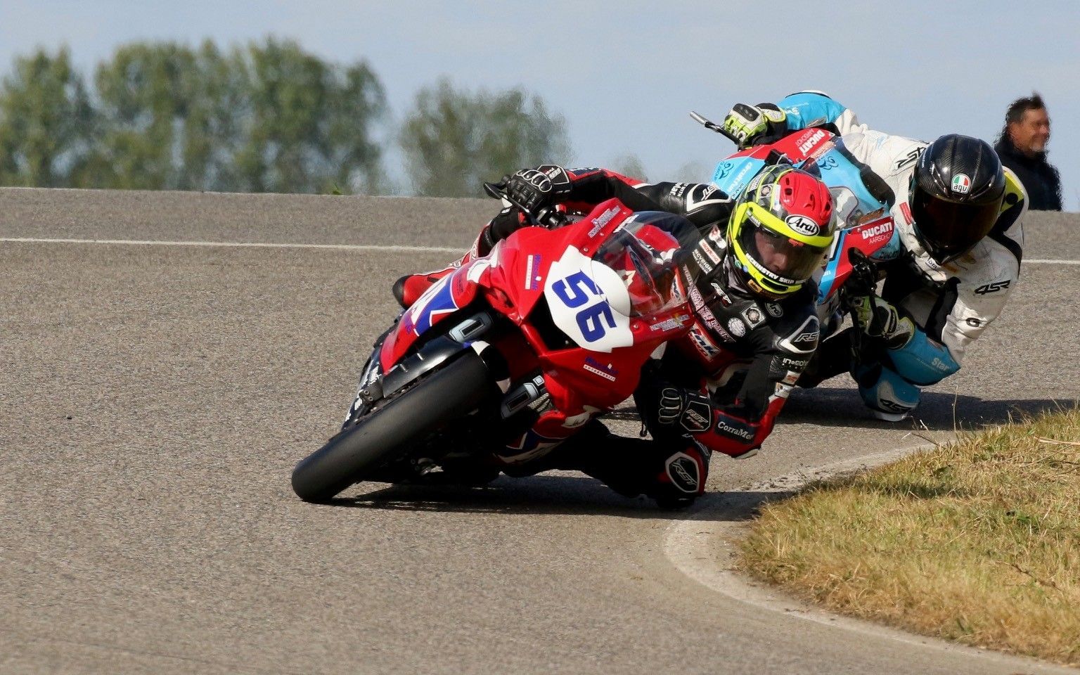 Twee motoren racen over een circuit, leunend in een bocht. Een rijder in rood en zwart rijdt voorop.
