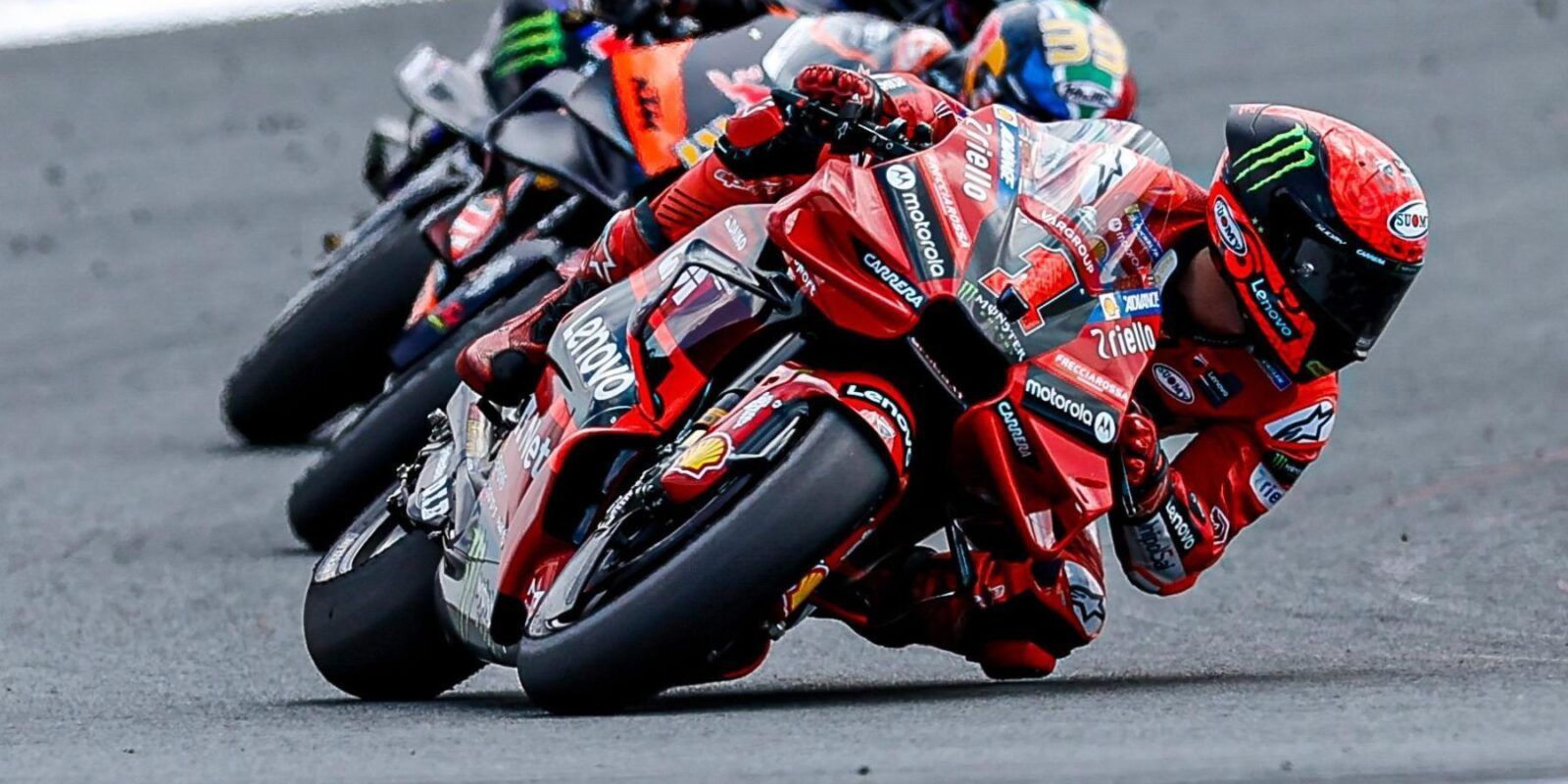 Motorrijders in rode racepakken buigen zich voorover in een bocht op een circuit en strijden hevig met andere motoren.