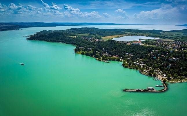Luchtfoto van het turquoise meer en de weelderig groene kustlijn met hier en daar gebouwen en een pier.