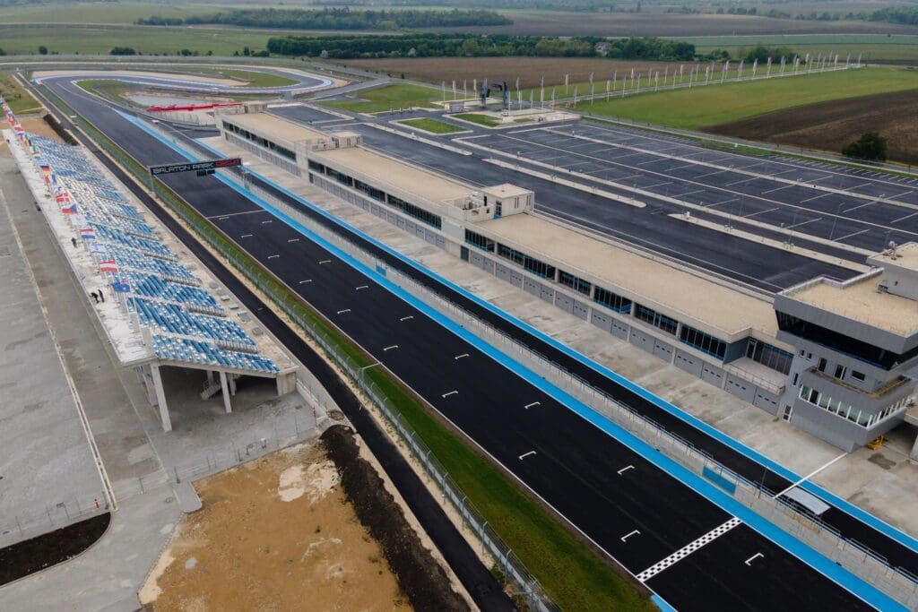 Luchtfoto van een renbaan met een lange zwarte asfaltbaan, blauwe hekken en tribunes met een gedeeltelijk overkapt dak.