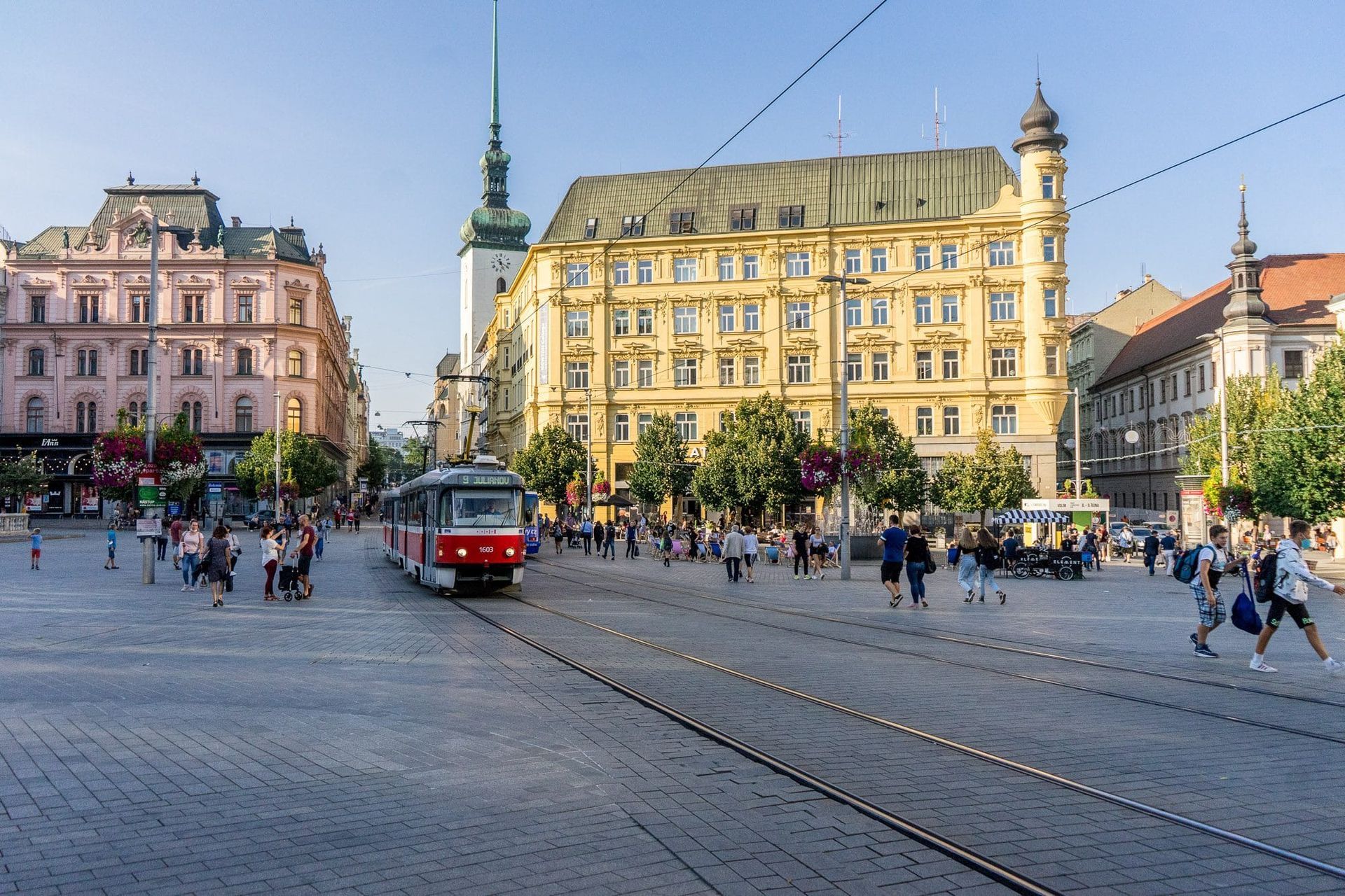 Een openbaar plein in Brno, Tsjechië, met een rode tram, voetgangers en historische gebouwen op de achtergrond onder een blauwe lucht.