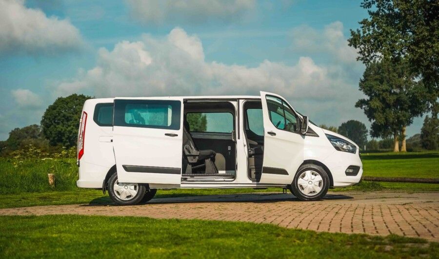 Witte Ford Transit-bus met open schuifdeur geparkeerd op een grasveld, blauwe lucht met wolken op de achtergrond.