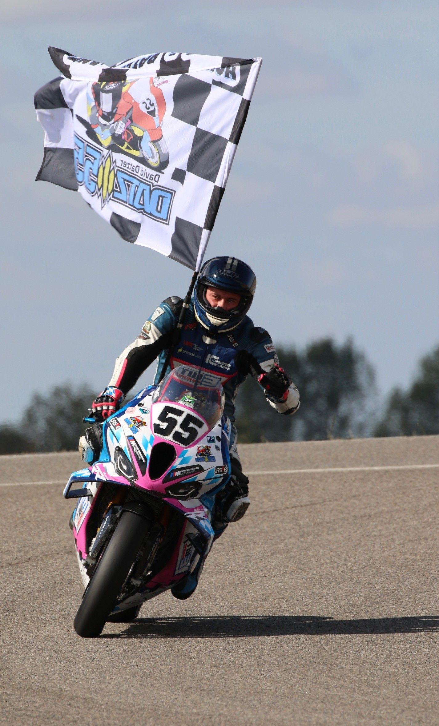 Motorcoureur op een roze en blauwe fiets, die op het circuit een geblokte vlag zwaait ter ere van zijn succes.