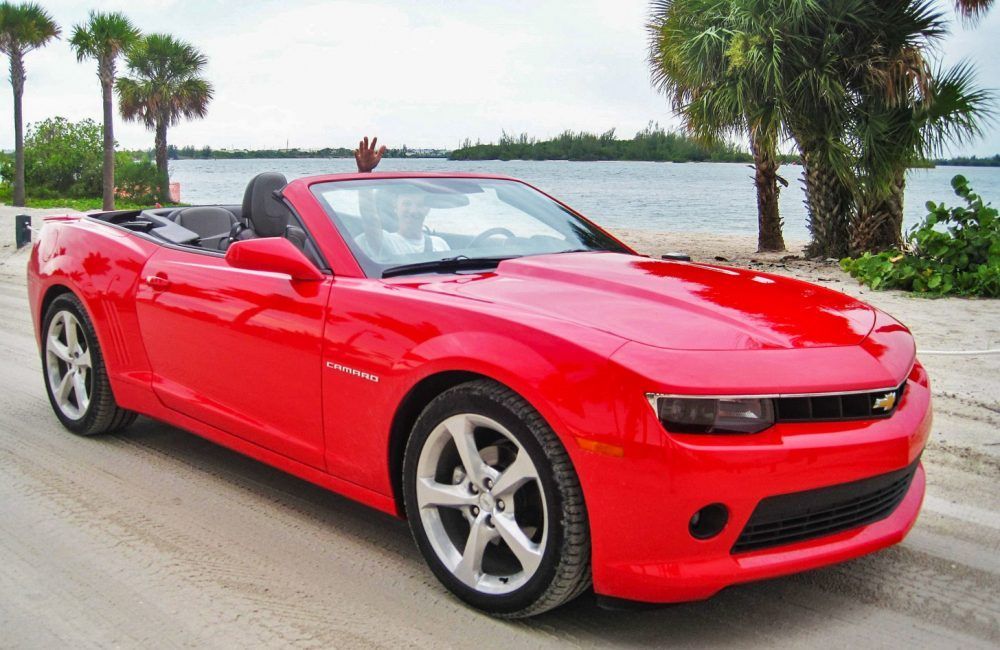 Rode Camaro cabriolet op een zandstrand, persoon zwaait vanaf de bestuurdersstoel, palmbomen en water op de achtergrond.