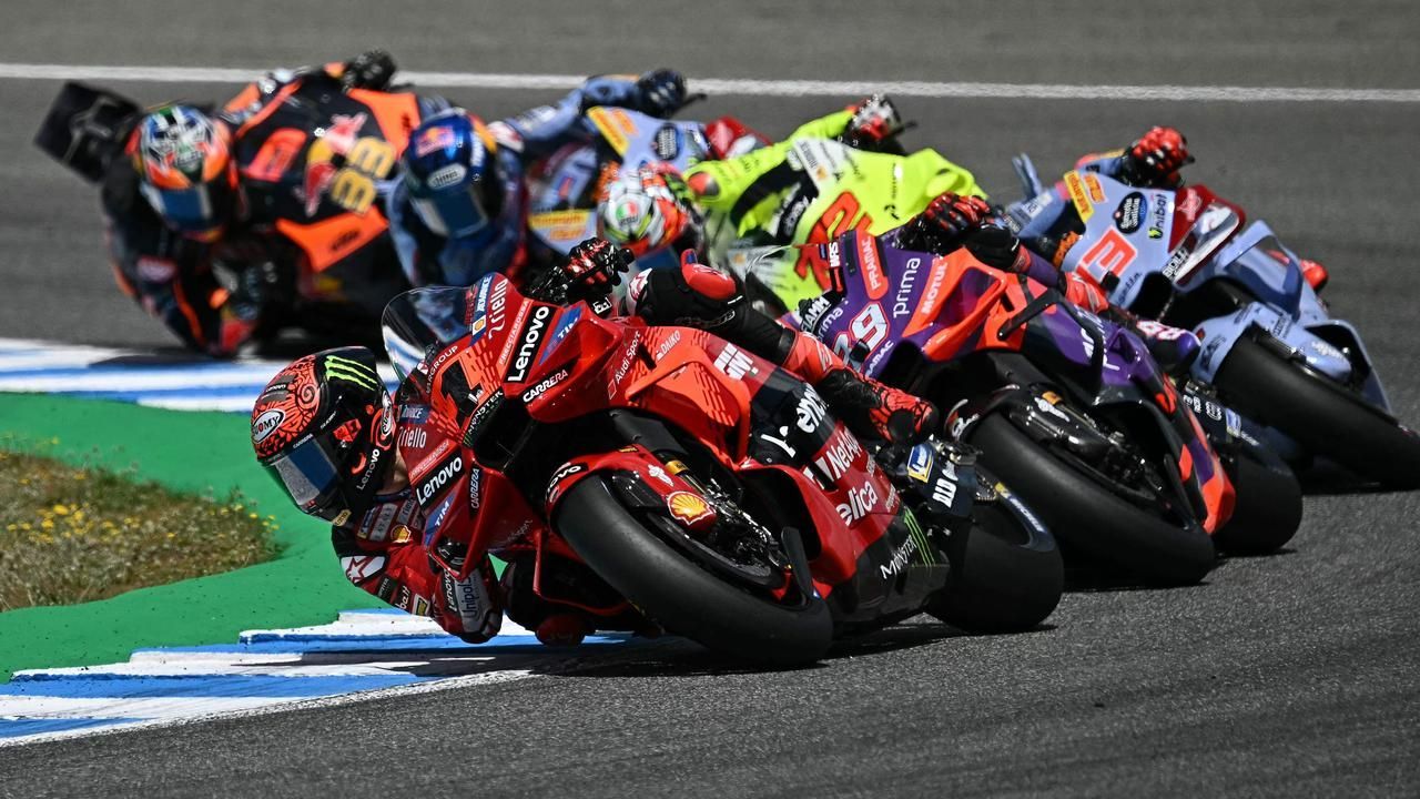 Motorcoureurs leunen dicht op elkaar in een bocht op een circuit. Rode, oranje en blauwe motoren zijn prominent aanwezig.