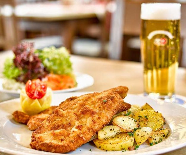 Bord met gepaneerde schnitzel met aardappelen, salade en een groot glas bier op een restauranttafel.