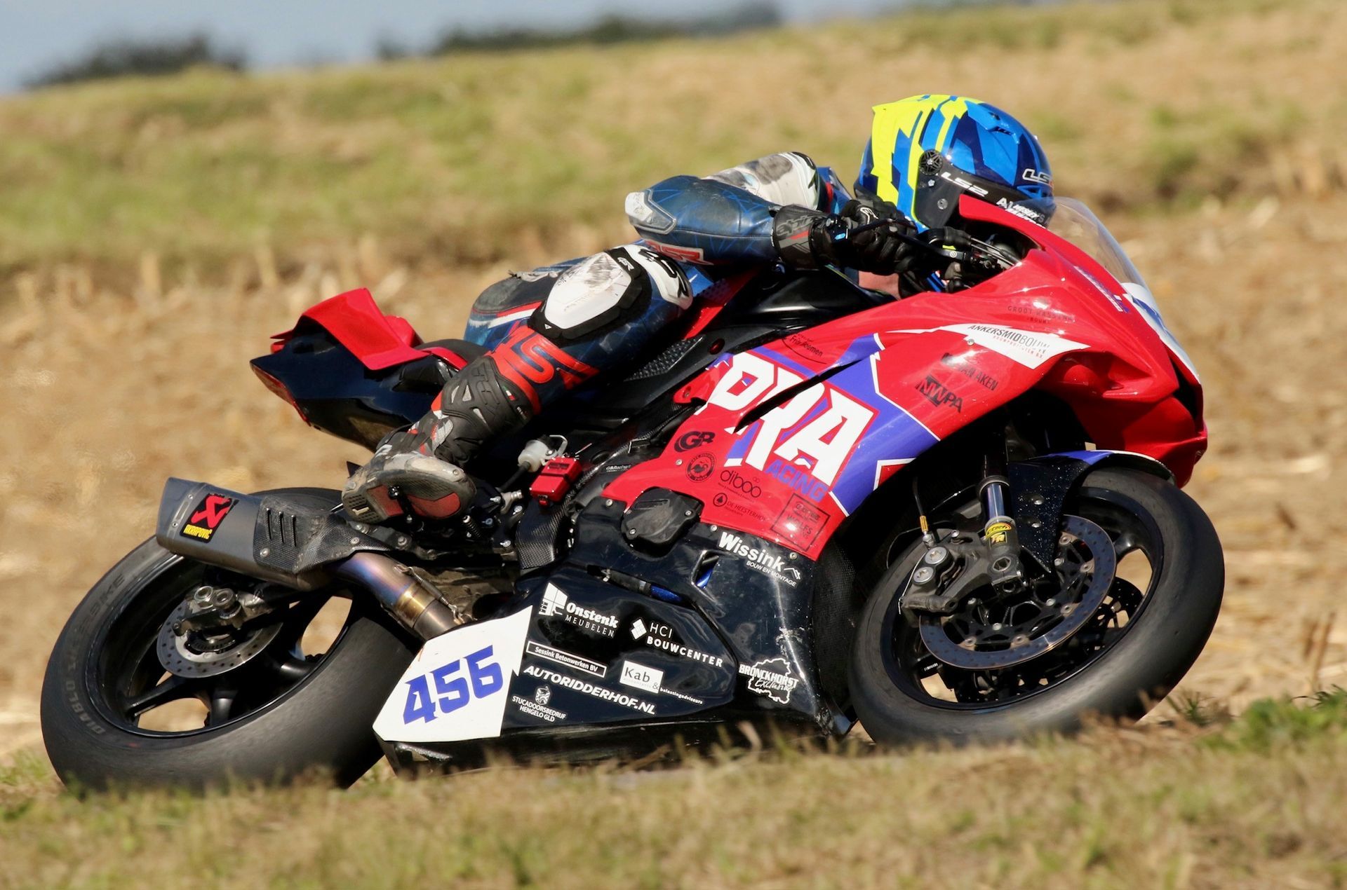 Motorcoureur in rood-blauw leer leunt in een bocht op een circuit. De motor is zwart-rood, nummer 456 is zichtbaar.