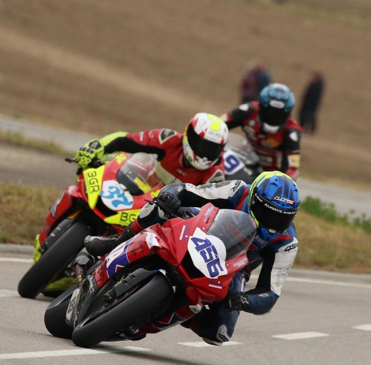 Motorcoureurs leunen in een bocht op een stratencircuit. Op de voorgrond zijn rood-wit-blauwe motoren en coureurs te zien.