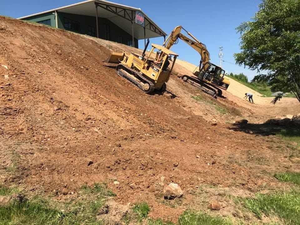 Two excavators working on a steep dirt slope near a building under a clear blue sky