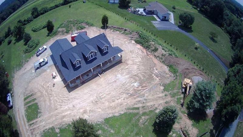 Aerial view of a house under construction on a dirt lot, surrounded by trees and a winding road.