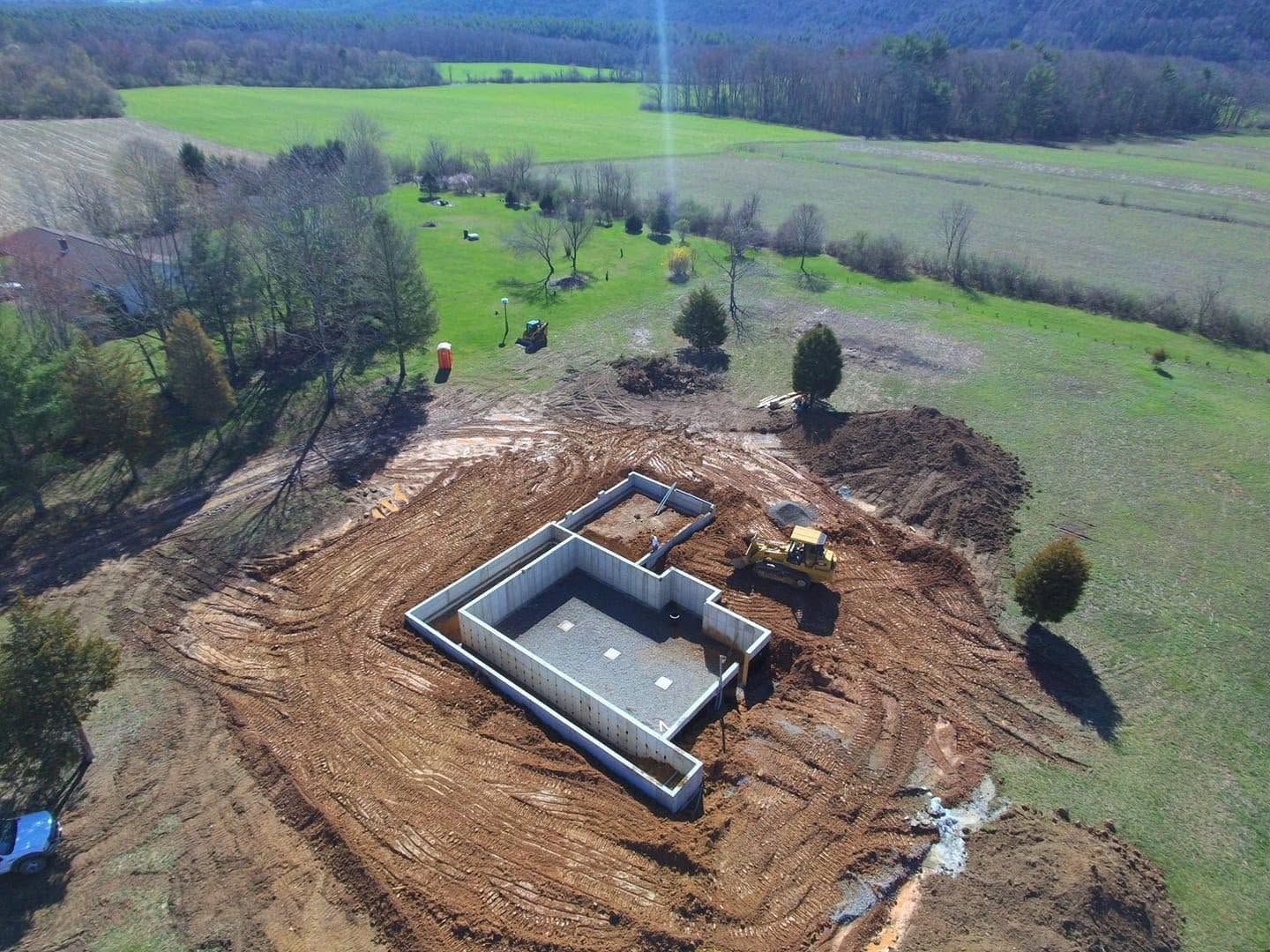 Aerial view of a concrete foundation under construction on a cleared wooded site with green fields nearby