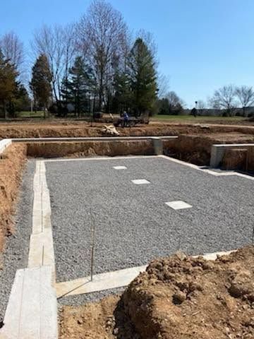 Gravel foundation area with concrete footings and rectangular cutouts in a wooded field under a clear sky