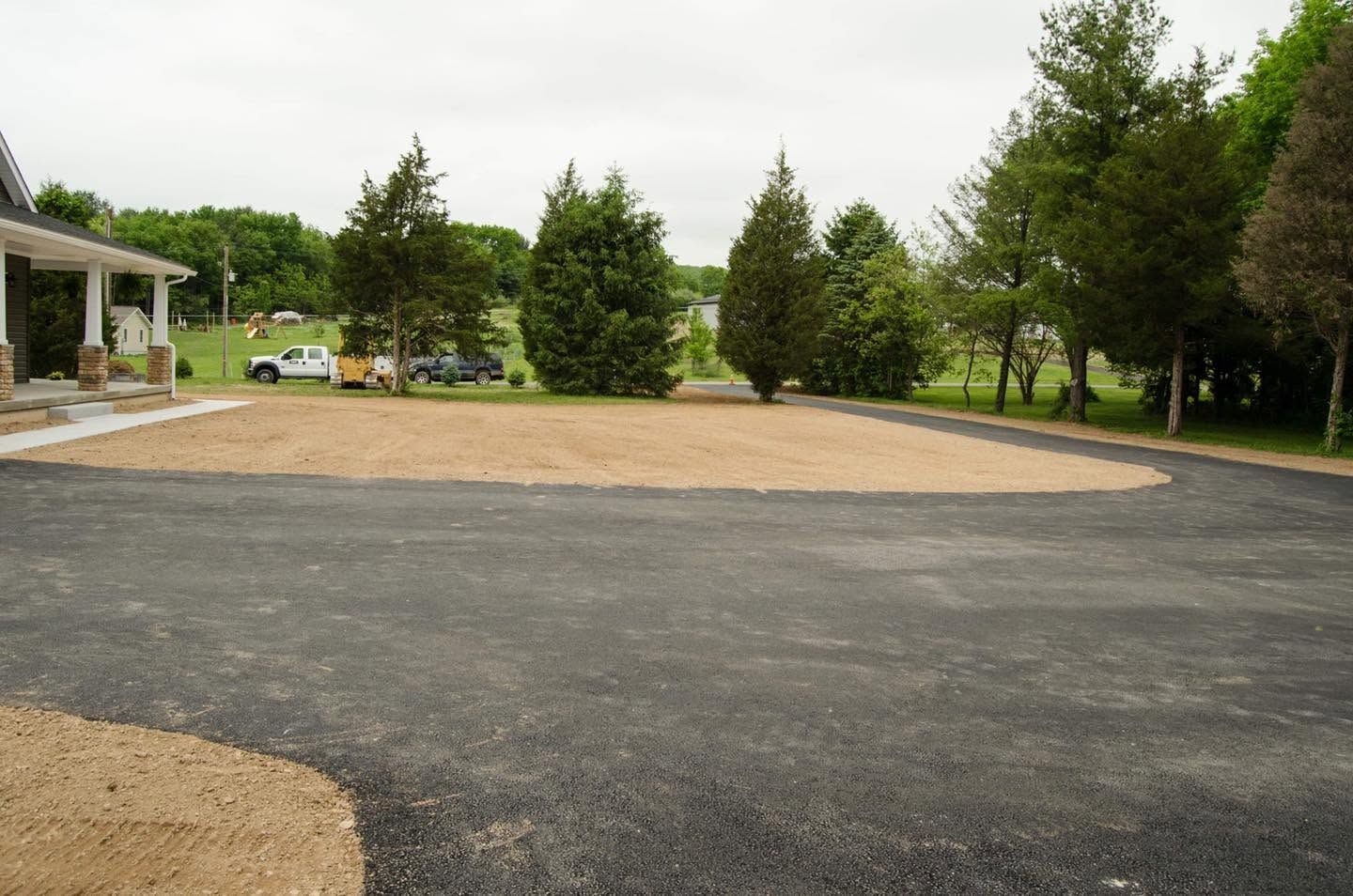 Gravel parking area beside a grassy lot with trees and a building on the left