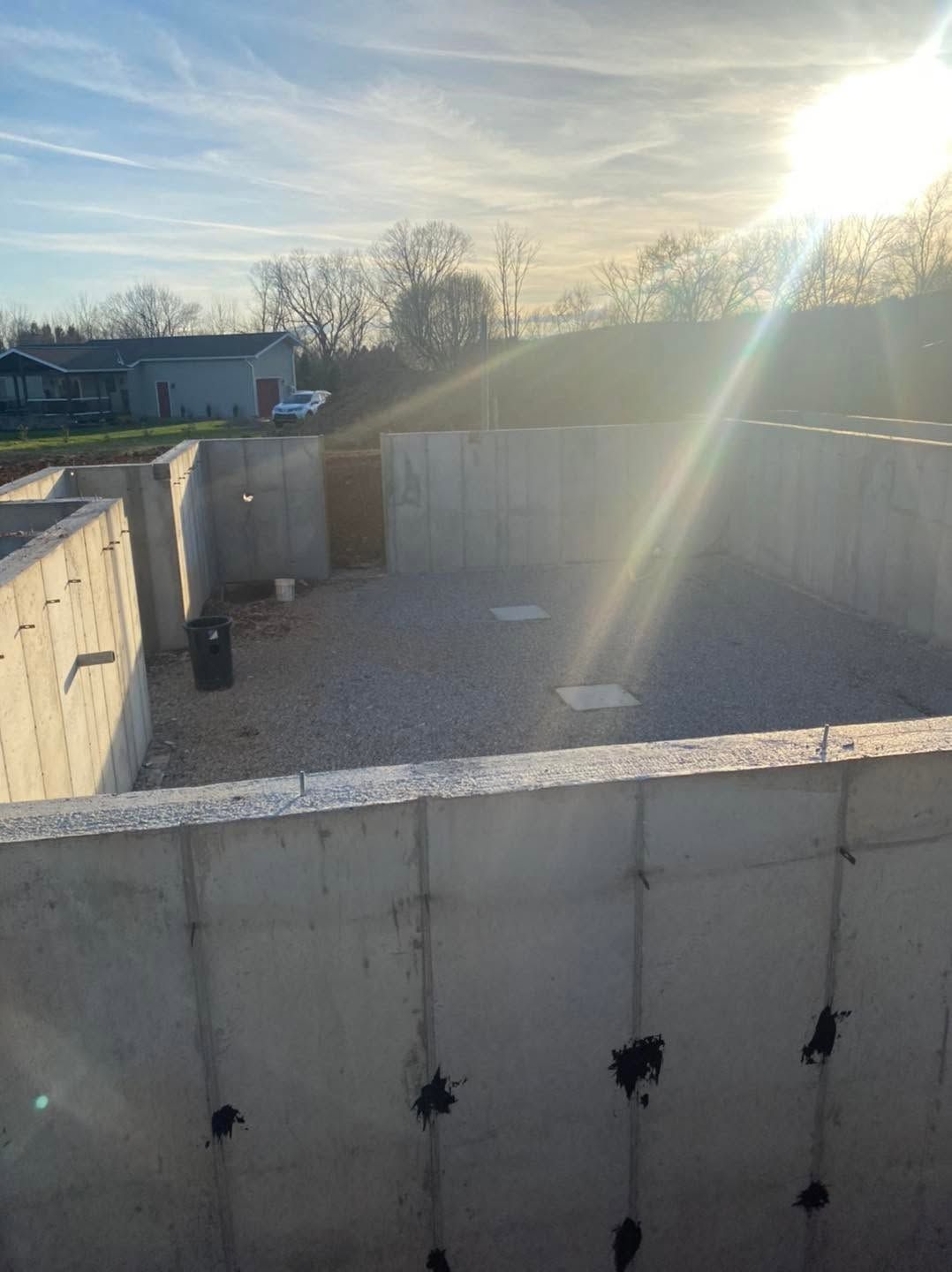 Sunlit concrete foundation walls and gravel lot under construction, with the sun low in the sky.