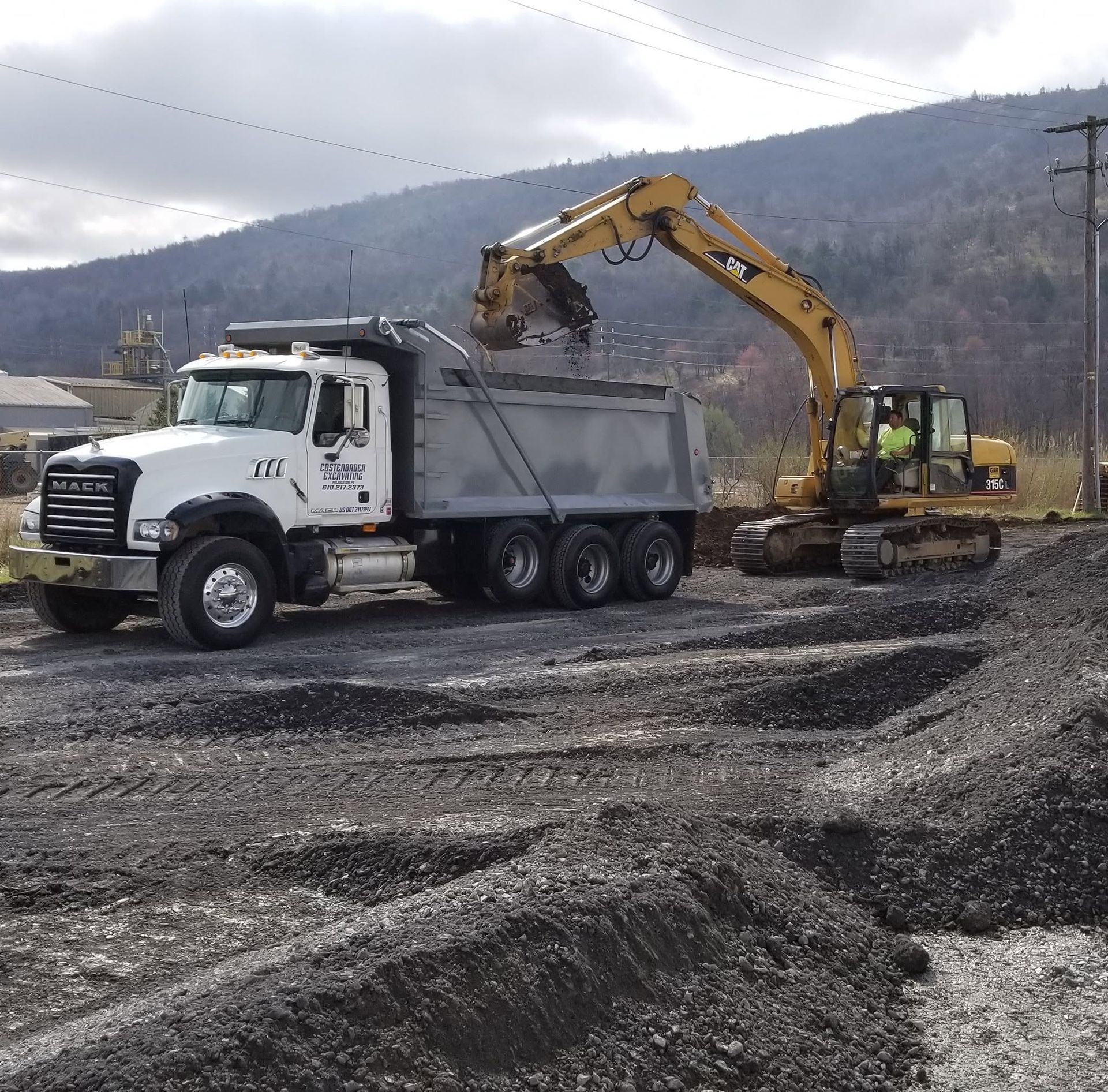 Excavator loading dirt into a white dump truck at a gravel construction site with mountains behind.
