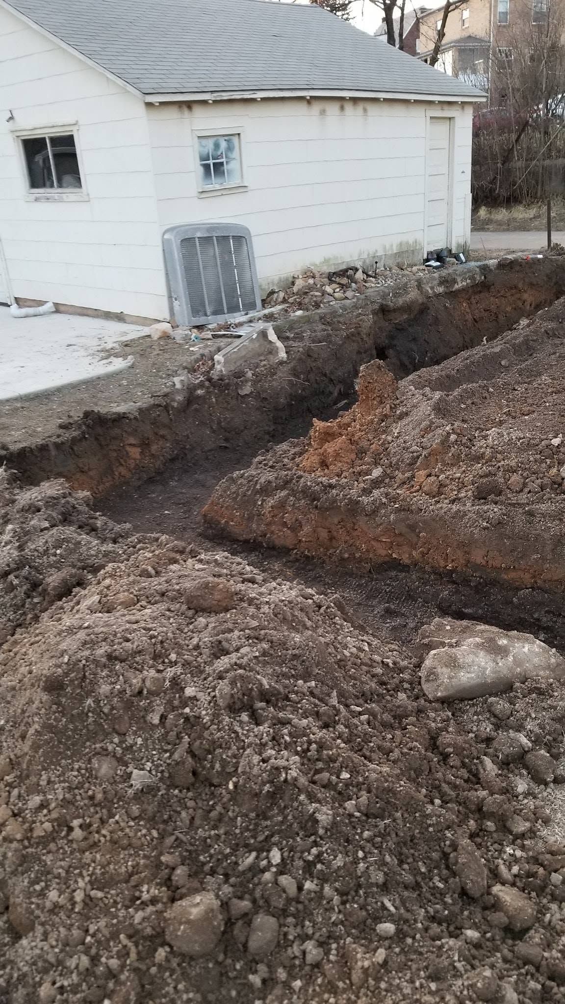 Trench dug beside a white house, with a pile of gravel and disturbed soil in the foreground.