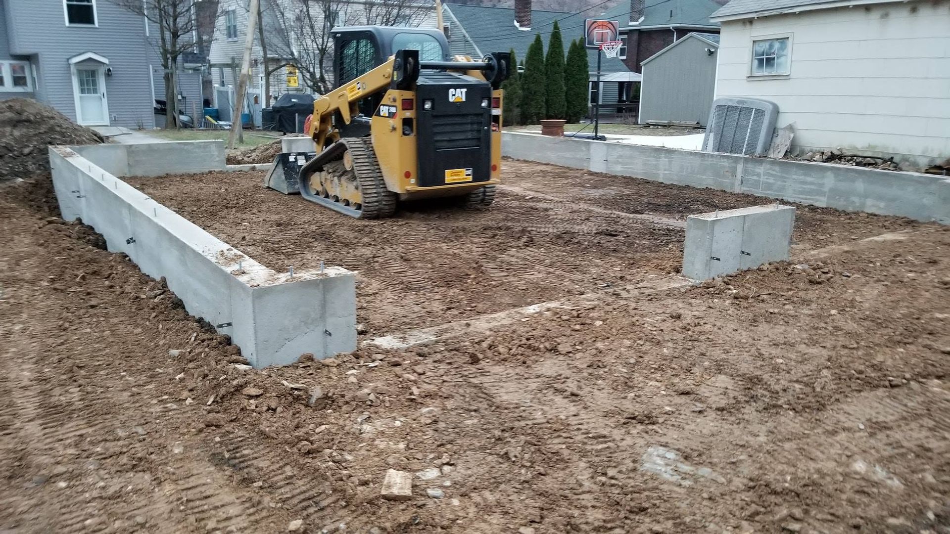 Small excavator grading a muddy foundation area bordered by concrete retaining walls in a residential yard.