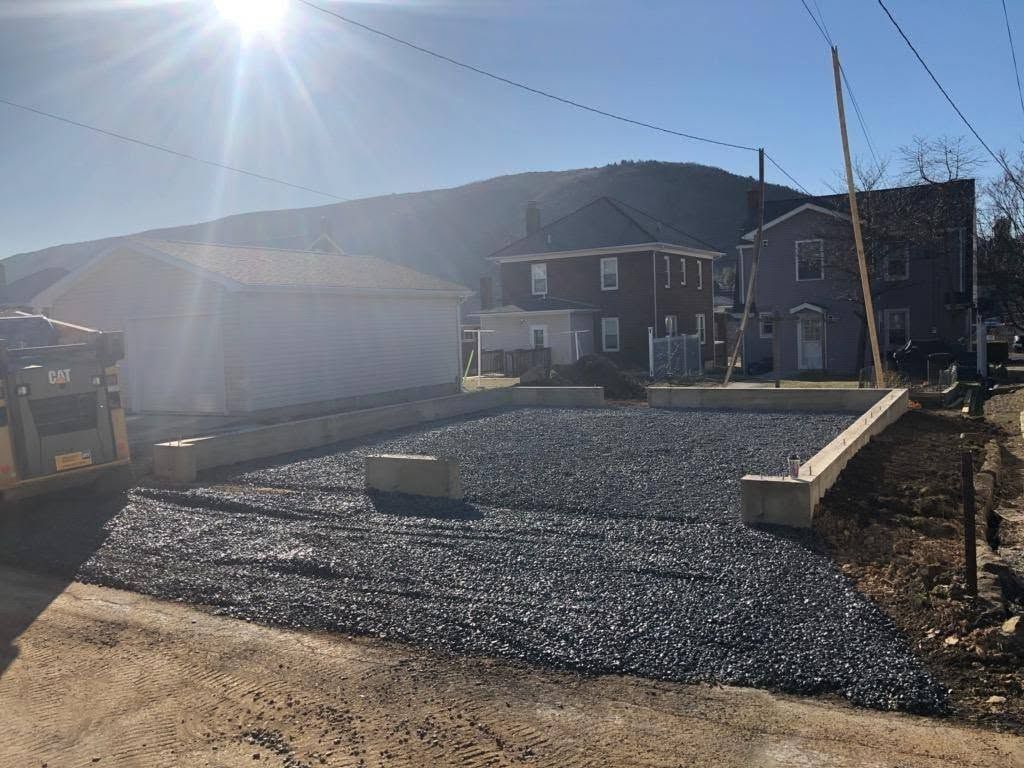 Gravel driveway under construction beside a house, with bright sun and mountains in the background.