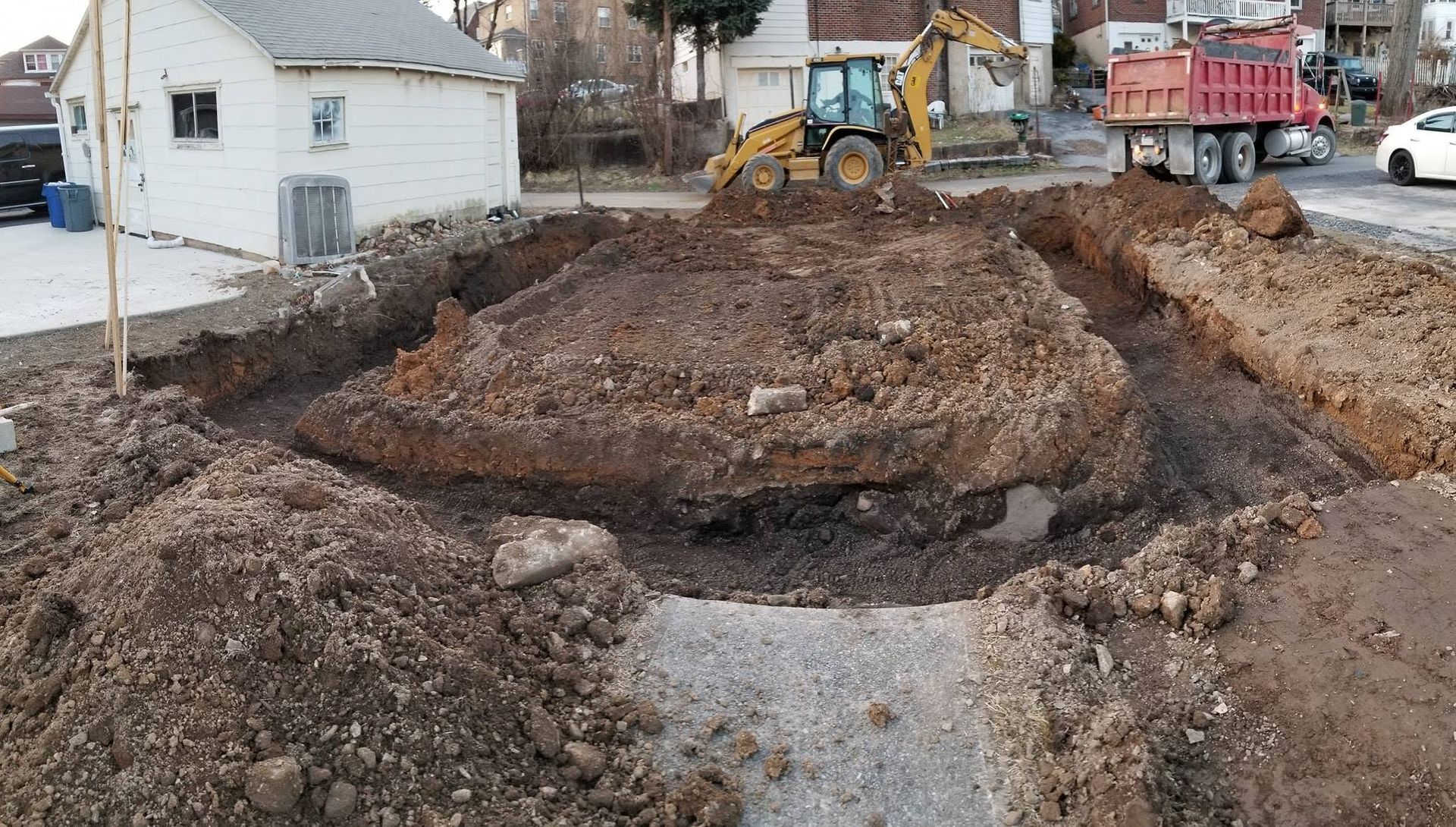 Construction site with excavator and dump trucks digging a muddy road trench near houses
